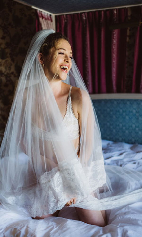 Braut in eleganter, weißer Hochzeitswäsche mit einem langen Schleier, die auf einem Bett sitzt und lacht. Die Szene wirkt verträumt und romantisch.