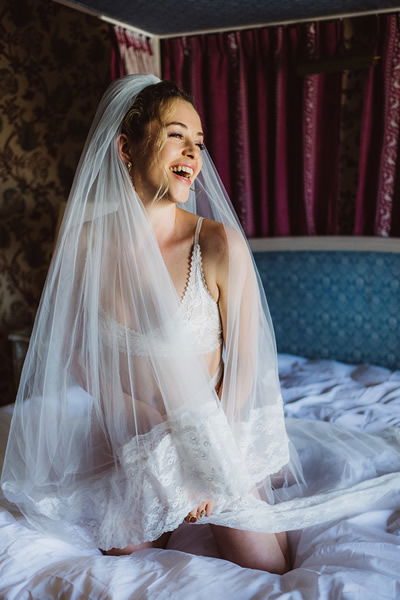 Braut in eleganter, weißer Hochzeitswäsche mit einem langen Schleier, die auf einem Bett sitzt und lacht. Die Szene wirkt verträumt und romantisch.