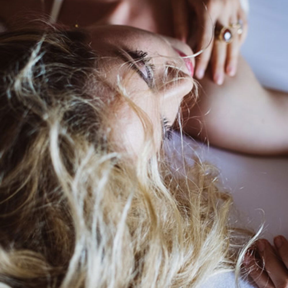 Intime Aufnahme einer Braut, die auf einem Bett liegt, mit welligem, blondem Haar. Ihre sanfte Pose und die ruhige Umgebung strahlen Gelassenheit und Eleganz aus.