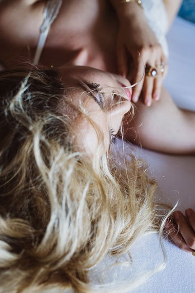 Intime Aufnahme einer Braut, die auf einem Bett liegt, mit welligem, blondem Haar. Ihre sanfte Pose und die ruhige Umgebung strahlen Gelassenheit und Eleganz aus.