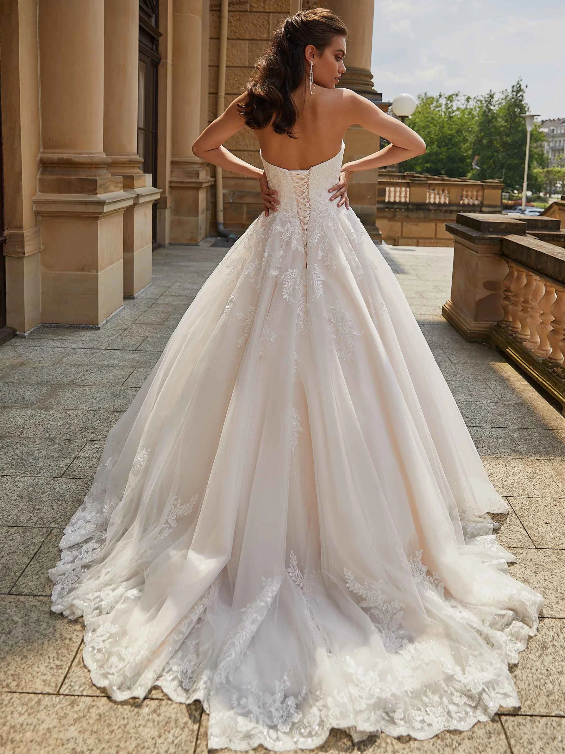 Braut in einem trägerlosen Hochzeitskleid mit romantischer Spitzenverzierung und langer Schleppe, die durch eine klassische Architekturkulisse geht.