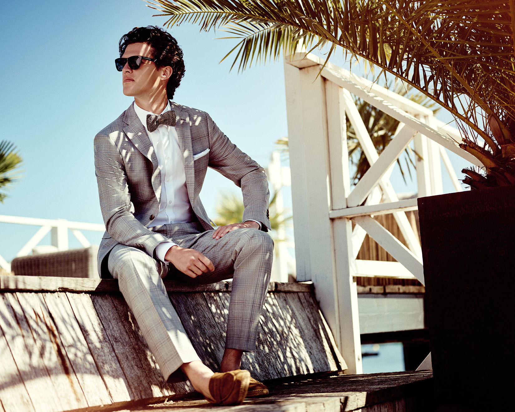 Junger Mann mit Sonnenbrille, grauem Anzug und Fliege lehnt sich entspannt zurück. Er sitzt an einem sonnigen Ort mit Palmen im Hintergrund.