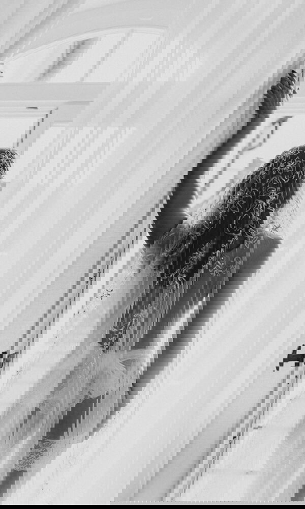 Eine Braut in verführerischer Hochzeitsdessous-Kombination mit langem Schleier steht an einer offenen Tür. Das schwarz-weiße Foto unterstreicht die romantische Stimmung.