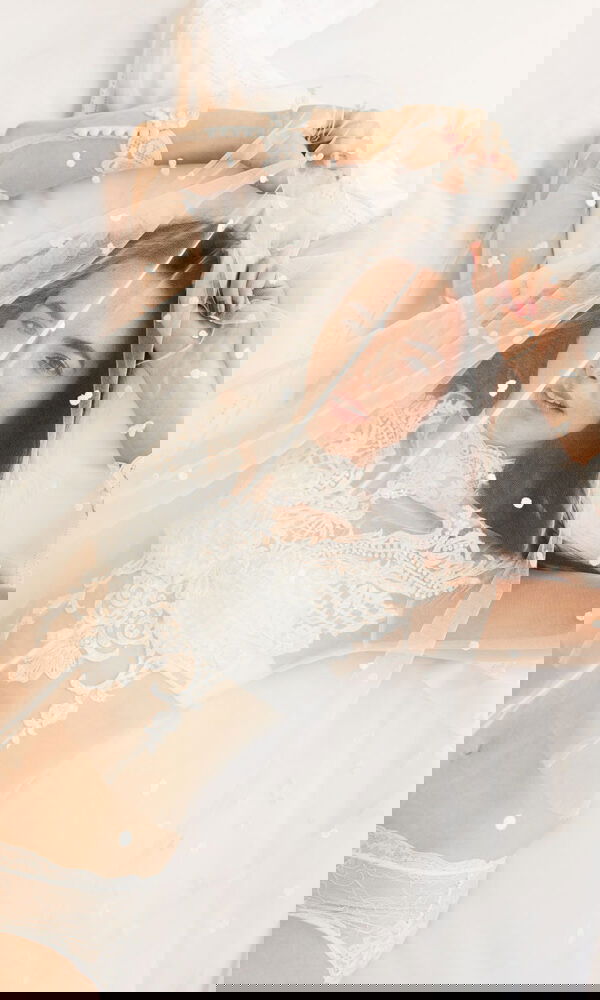 Ein intimer Moment: Eine Braut liegt auf dem Bett und spielt mit ihrem transparenten Schleier. Die edle Spitze ihrer Hochzeitslingerie ergänzt den verträumten Look.