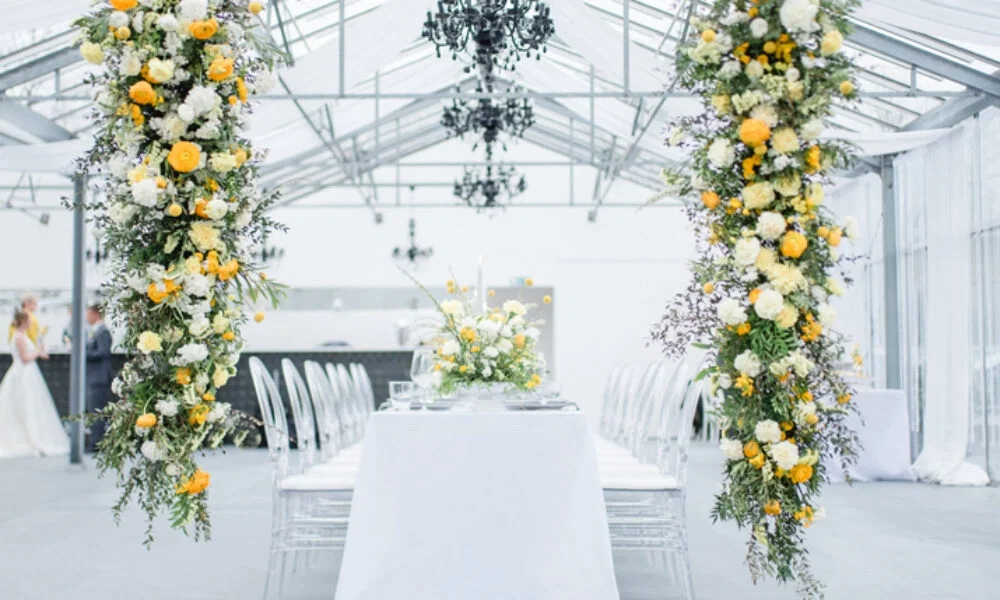Romantische Hochzeitsdekoration in einem Gewächshaus mit floralen Säulen in Gelb und Weiß, einem eleganten Tischarrangement und luxuriösen Kristallleuchtern.