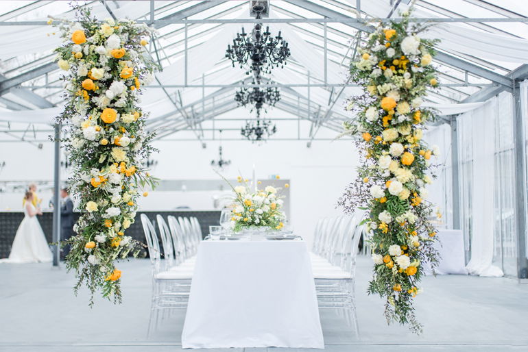 Moderner Hochzeitstisch, umgeben von gelb-weißen Blumeninstallationen in einem Gewächshaus, mit kunstvollen Kronleuchtern als Teil der Dekoration.