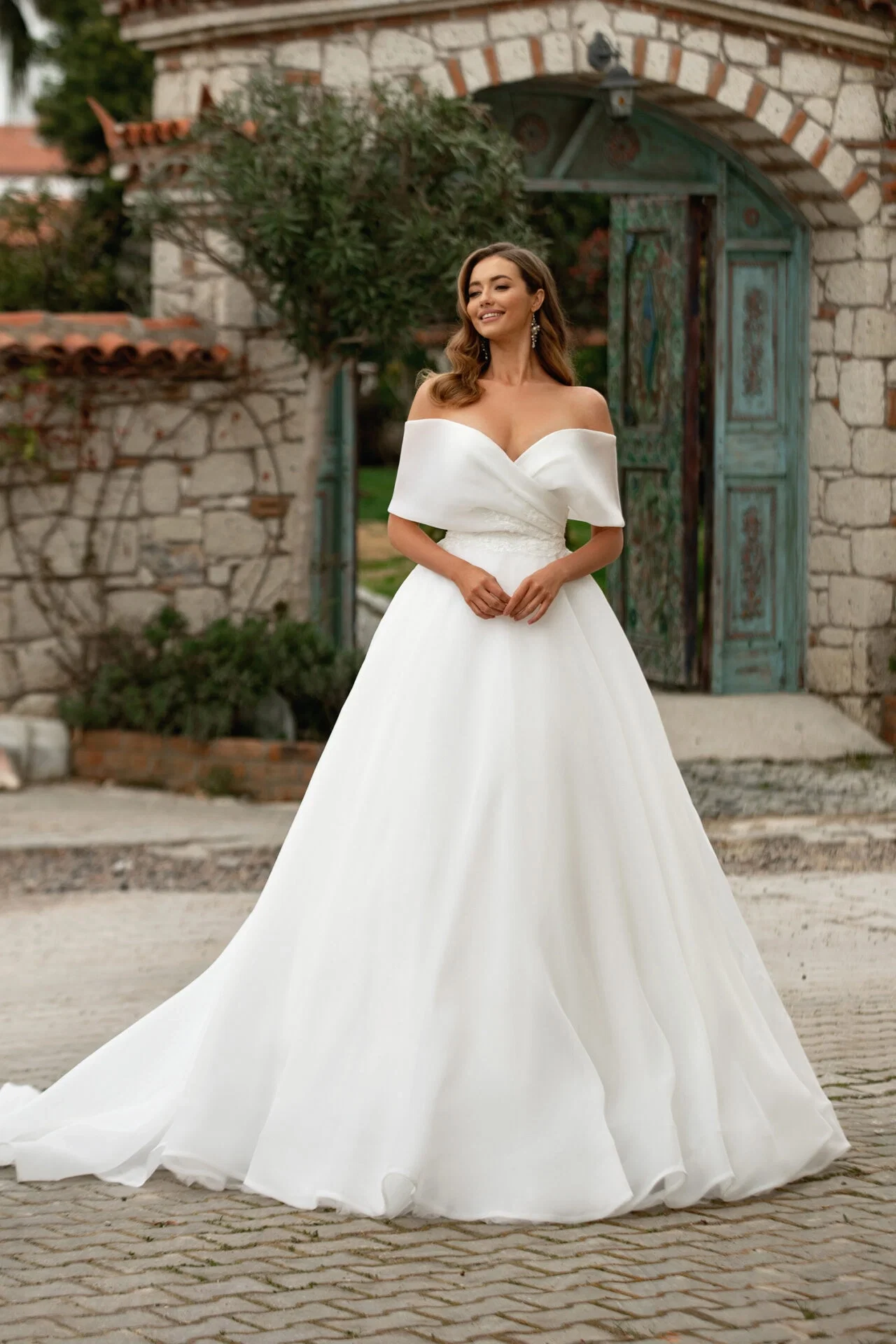 Braut in einem klassischen weißen Ballkleid mit schulterfreiem Ausschnitt, posierend vor einer charmanten Steinmauer und einem antiken Tor.