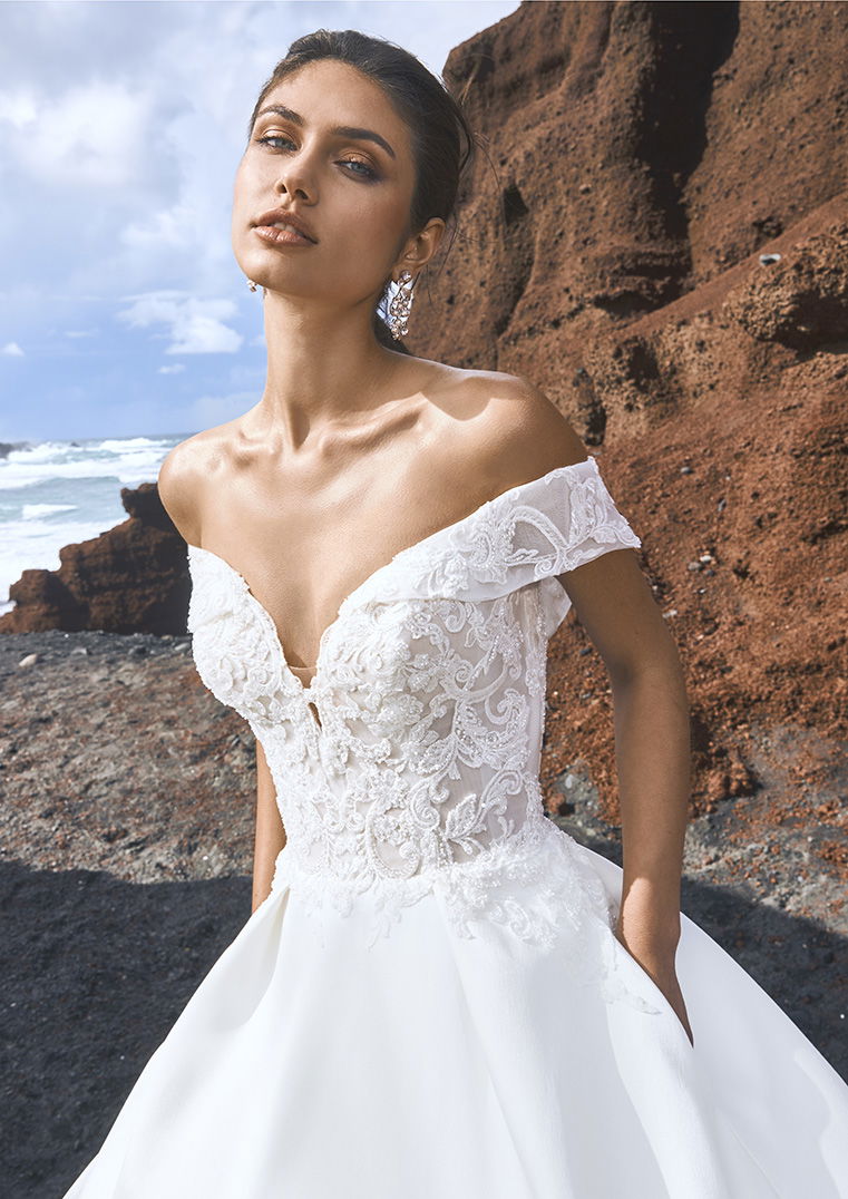 Elegante Braut in einem spitzenverzierten, schulterfreien Kleid, posierend vor rot-braunen Felsen und einem Meereshorizont im Hintergrund.