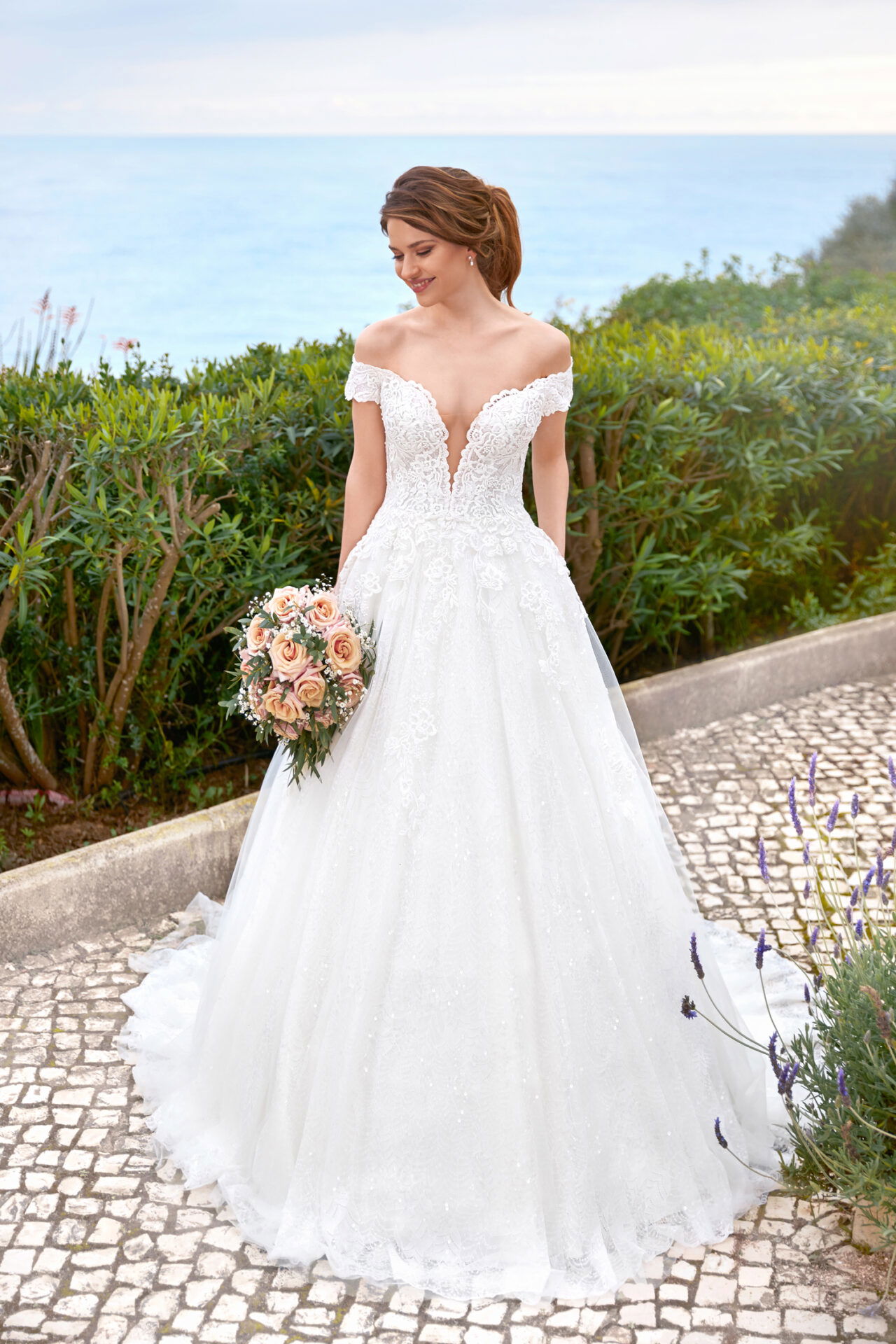 Elegante Braut in einem Spitzen-Brautkleid mit tiefem Ausschnitt, posierend mit Blumenstrauß auf einer gepflasterten Terrasse mit Meerblick.