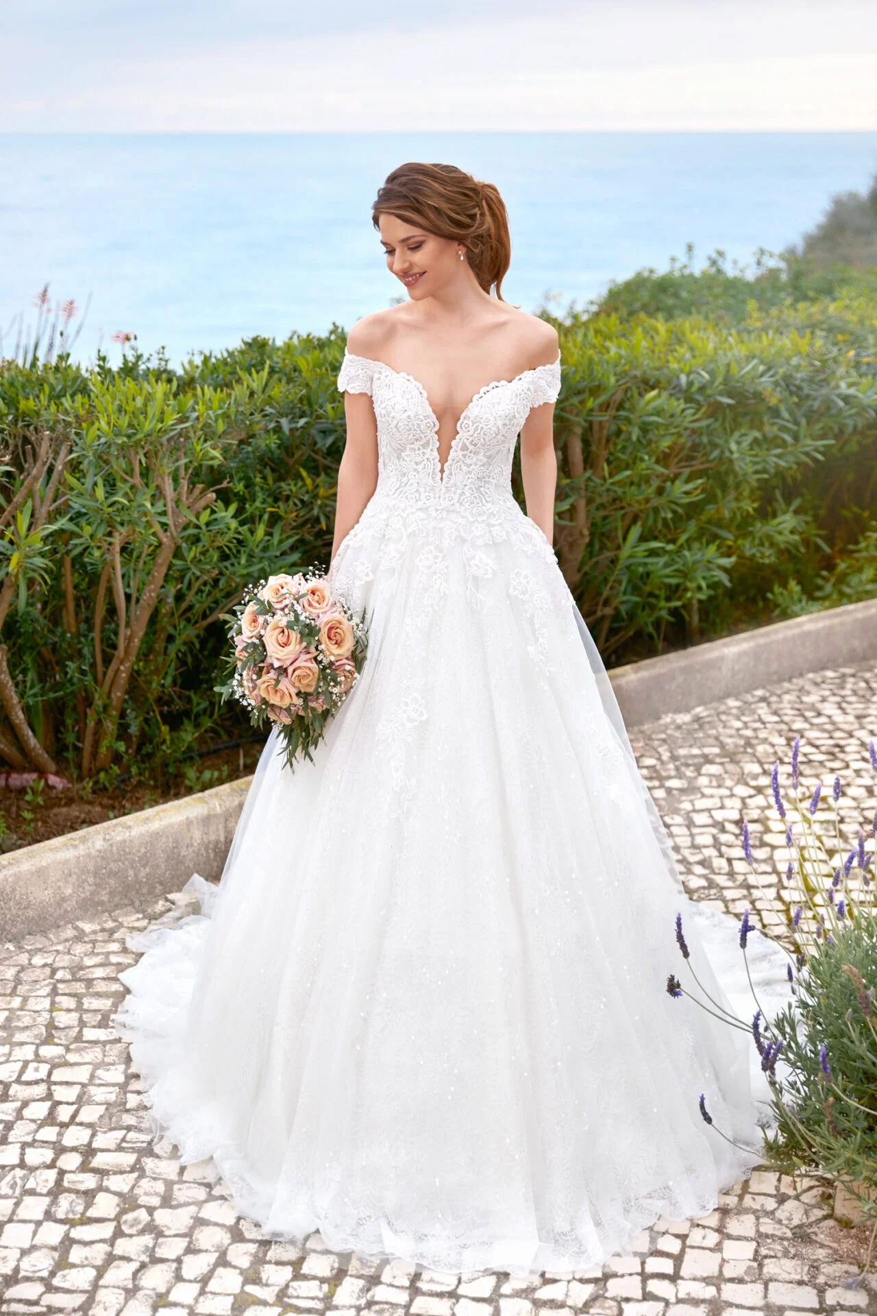 Elegante Braut in einem Spitzen-Brautkleid mit tiefem Ausschnitt, posierend mit Blumenstrauß auf einer gepflasterten Terrasse mit Meerblick.