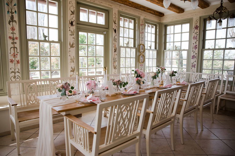 Stilvoll eingedeckter Hochzeitstisch in einem sonnigen Raum, umgeben von weißen Holzstühlen, Blumenvasen und sanftem Tageslicht durch große Fenster.