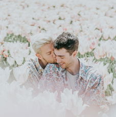 Ein junges Paar in floralen Hemden sitzt eng umschlungen in einem Meer aus Tulpen. Einer der Männer lehnt sich nach vorn und küsst seinen Partner liebevoll auf die Wange, während dieser mit einem sanften Lächeln die Augen schließt. Die Umgebung ist in sanfte Pastellfarben getaucht.