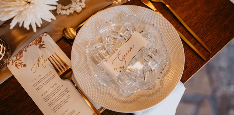 Festliche Tischdekoration mit goldenen Akzenten, Kristallgeschirr und einer Menükarte, die stilvoll auf den Hochzeitstisch des Bräutigams platziert wurde.
