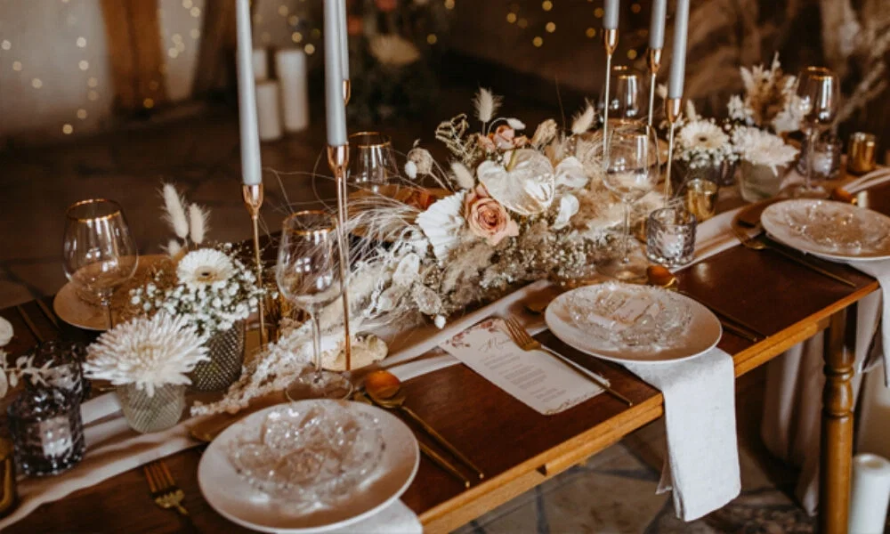 Stimmungsvolle Hochzeitsdekoration auf einem Holztisch mit klaren Gläsern, hellen Stoffservietten und kunstvollen Blumengestecken in Creme- und Rosétönen, ergänzt durch Kerzenschein.