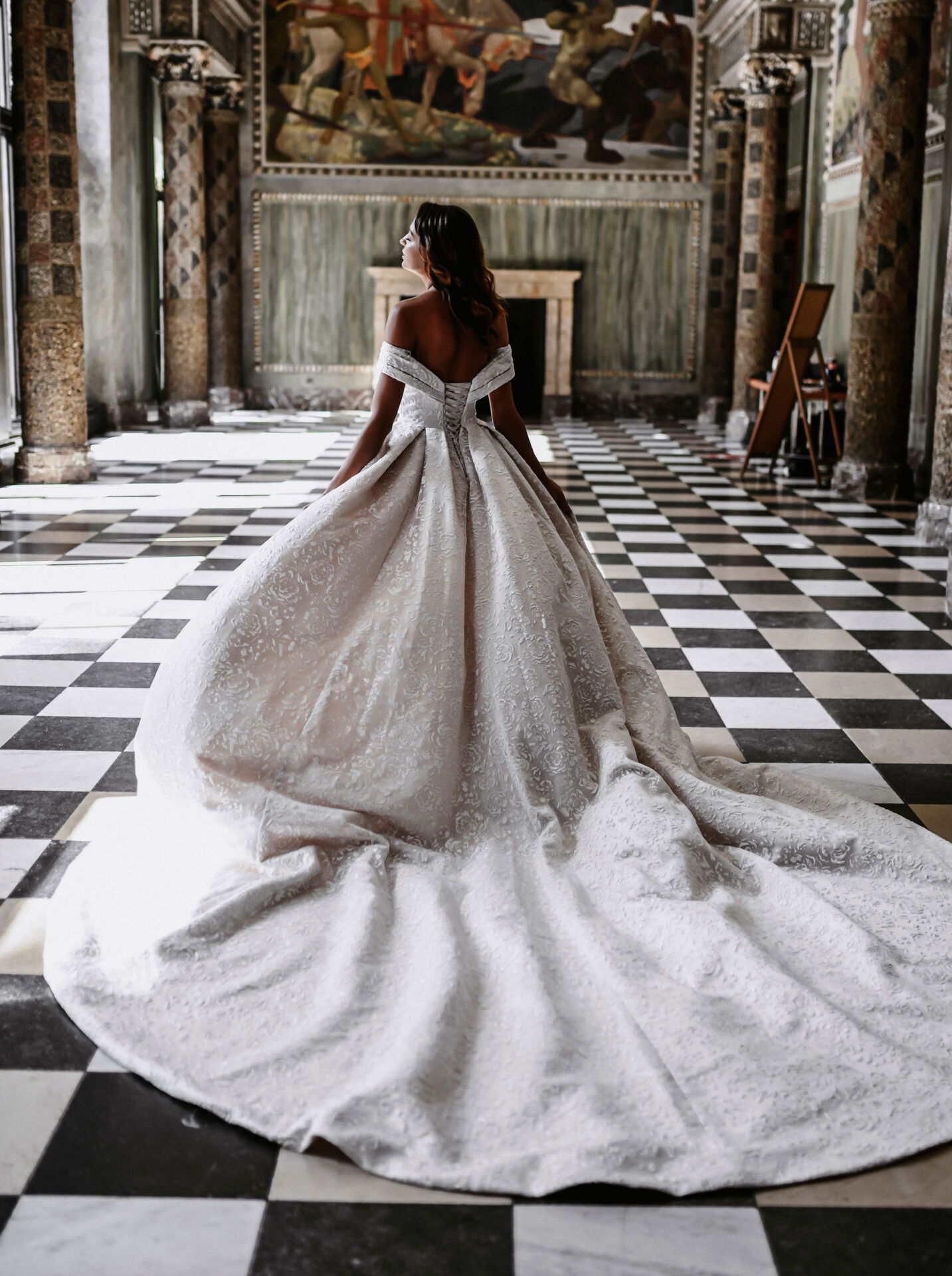 Braut trägt ein aufwendiges, schulterfreies Hochzeitskleid mit langer Schleppe und posiert in einem klassischen Saal mit schwarz-weißem Boden.
