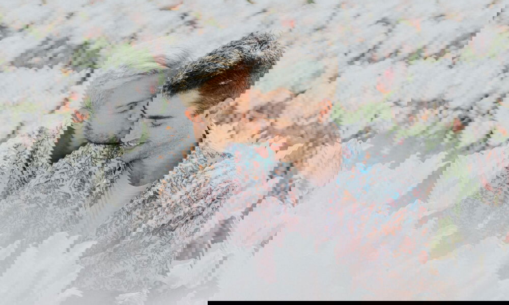 Ein junges Paar sitzt eng umschlungen zwischen blühenden Tulpen. Während einer der Männer liebevoll den Kopf seines Partners küsst, genießt dieser den Moment mit einem Lächeln. Ihre abgestimmten Hemden mit floralen Mustern ergänzen die sanfte, romantische Atmosphäre des Bildes.