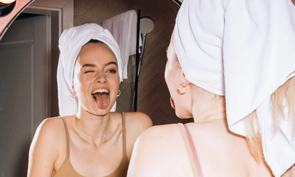 Vor einem Spiegel steht eine Frau mit Handtuchturban, die mit einem humorvollen Gesichtsausdruck ihre Zunge zeigt. Die entspannte Atmosphäre des Badezimmers deutet auf eine entspannte Pflegeroutine hin.