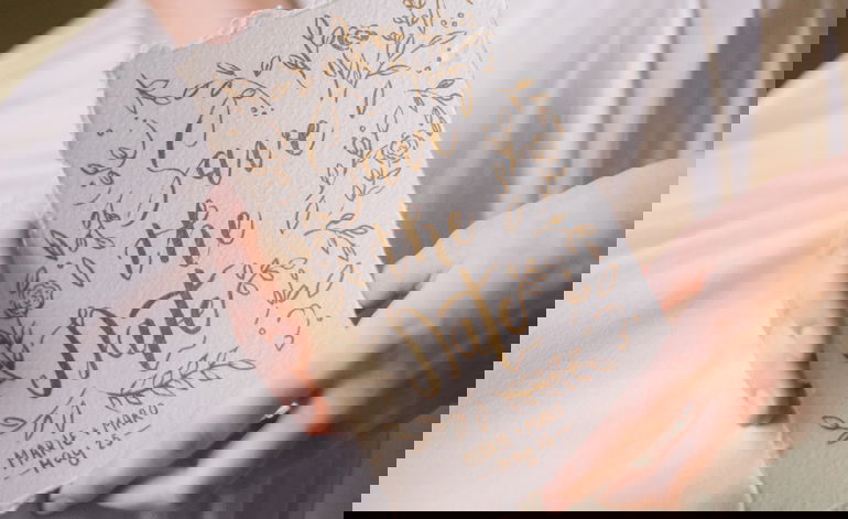 Handgeschriebene Save-the-Date-Karte mit goldenen Blumenornamenten auf strukturiertem Papier, gehalten von einer Braut in einem weißen Kleid.