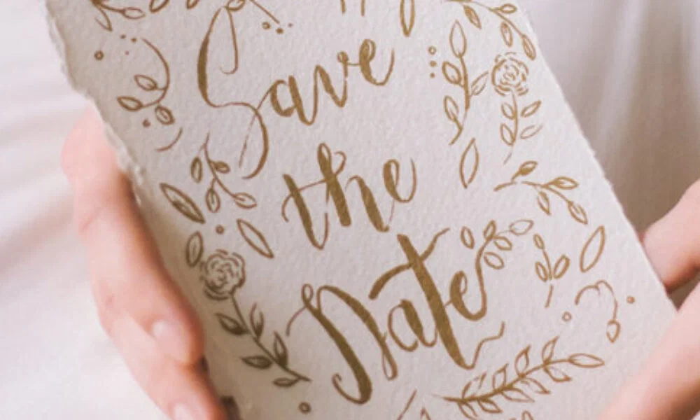 Elegante Save-the-Date-Karte mit floralem Muster in Gold, handgeschrieben und von einer Person in Nahaufnahme präsentiert.