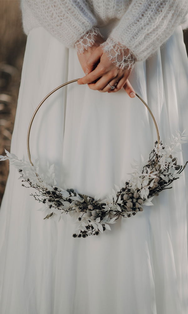 Die Hände einer Braut ruhen auf einem filigranen, mit Trockenblumen dekorierten Metallring. Sie trägt einen weichen Strickpullover über ihrem Brautkleid, was dem Bild eine gemütliche, natürliche Atmosphäre verleiht.