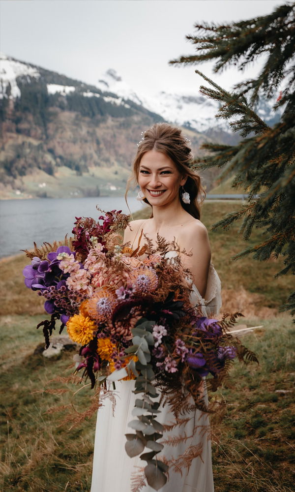 Eine junge Frau im weißen Brautkleid posiert mit einem riesigen, kunstvoll gebundenen Blumenstrauß in satten, warmen Farben. Sie steht in einer alpinen Umgebung mit Nadelbäumen, grünen Wiesen und einem klaren Bergsee.