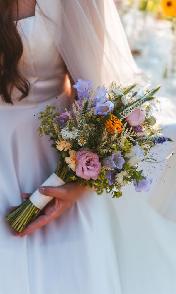 Eine Braut mit langem, leicht gewelltem Haar trägt einen klassischen Schleier und hält einen kunstvoll arrangierten Blumenstrauß mit blauen, rosa und gelben Akzenten, während sie sanft die Stiele umschließt.