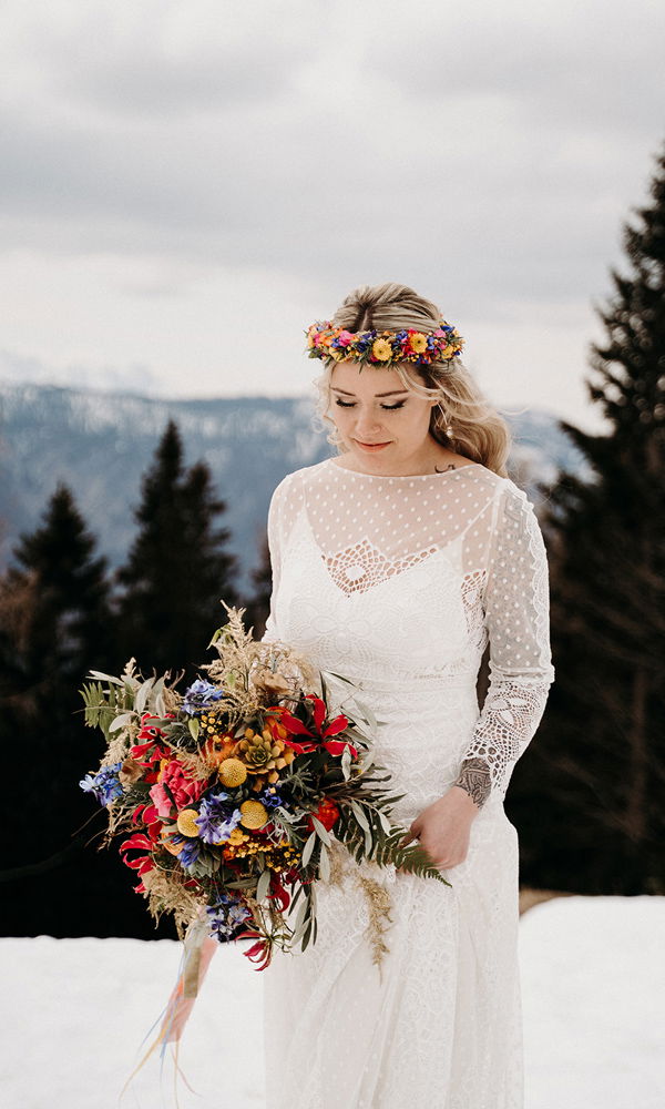 Vor einer malerischen Berglandschaft steht eine Braut im Boho-Stil mit einem romantischen Spitzenkleid. Sie hält einen natürlichen Blumenstrauß mit wilden roten, blauen und gelben Blüten in den Händen.