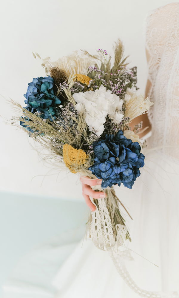 Ein handgebundener Trockenblumenstrauß in Blau, Weiß und Gelb wird von einer Braut mit Spitze und Tüll in der Hand gehalten. Die filigranen Details des Straußes unterstreichen den Vintage-Charakter.