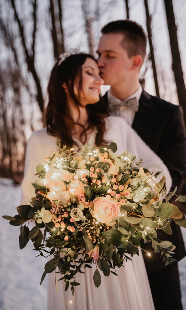 Eine verliebte Braut mit einem verträumten Lächeln lehnt sich an ihren Bräutigam, während er sie liebevoll küsst. Ihr blütenreicher Strauß mit sanftem Lichtschein verleiht dem Bild eine magische Atmosphäre.
