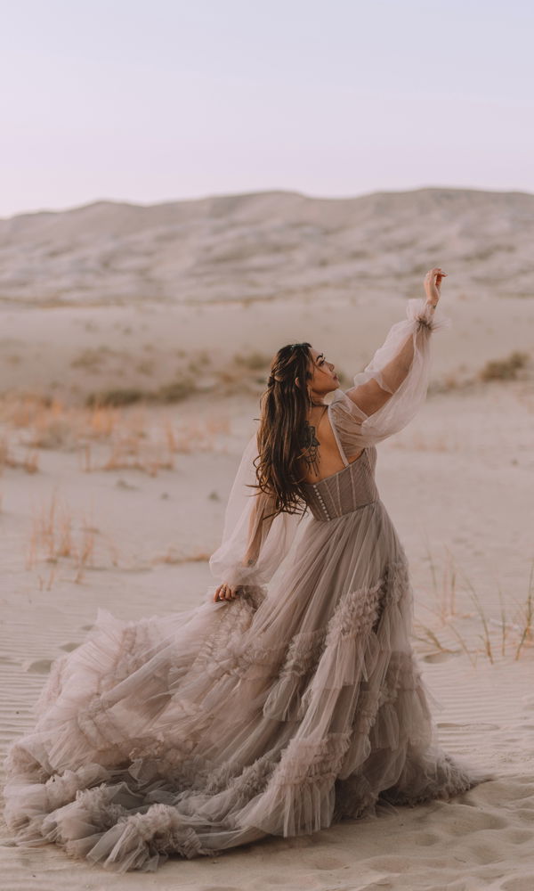 Traumhafte Braut im voluminösen, mehrlagigen Boho-Kleid mit zarten Ärmeln, vor der Kulisse einer sanften Wüste.