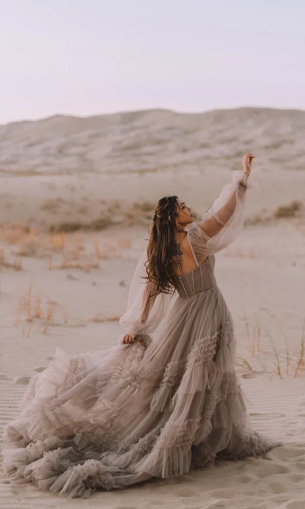Traumhafte Braut im voluminösen, mehrlagigen Boho-Kleid mit zarten Ärmeln, vor der Kulisse einer sanften Wüste.