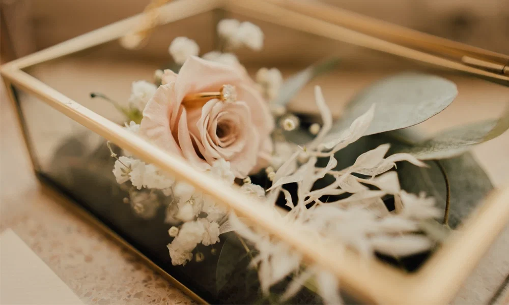 Eine dekorative Glasbox mit goldenen Akzenten, gefüllt mit einer rosafarbenen Rose, weißen Blüten und zartem Eukalyptus – eine stilvolle Ergänzung für jede Hochzeit.