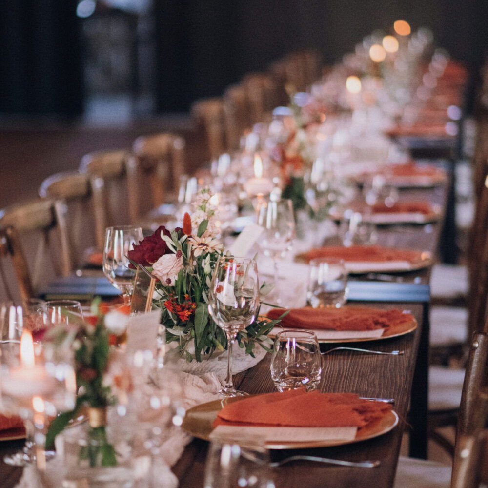 Der gedeckte Hochzeitstisch mit rustikalem Holzdesign und sanften Orangetönen wird durch Blumen und zartes Kerzenlicht romantisch beleuchtet.