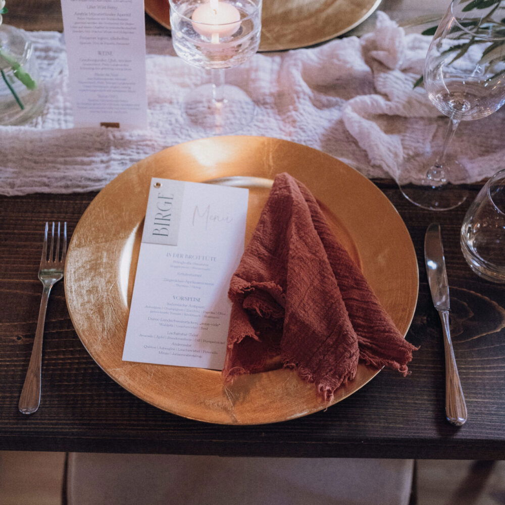 Ein festlich gedeckter Tisch mit goldenen Tellern, orangefarbenen Servietten und stilvoller Blumendekoration aus roten und weißen Blüten. Eine brennende Kerze im Glas fügt ein warmes, romantisches Licht hinzu.