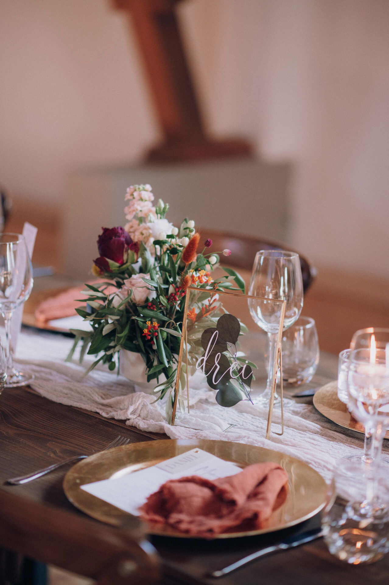 Eine elegante Hochzeitstafel, dekoriert mit Blumen, Weingläsern und einem Namensschild mit der Aufschrift „Drei“. Die Tischdeko ist in warmen Tönen gehalten, mit goldenen Tellern und rustikalen Servietten.
