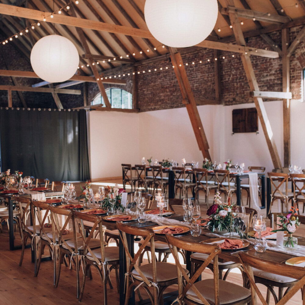 Ein rustikaler Hochzeitssaal mit Holzbalken, langen Tafeln und strahlend weißen Papierlaternen an der Decke. Die Tische sind für das Hochzeitsessen vorbereitet und mit eleganten Gläsern und Blumen geschmückt.