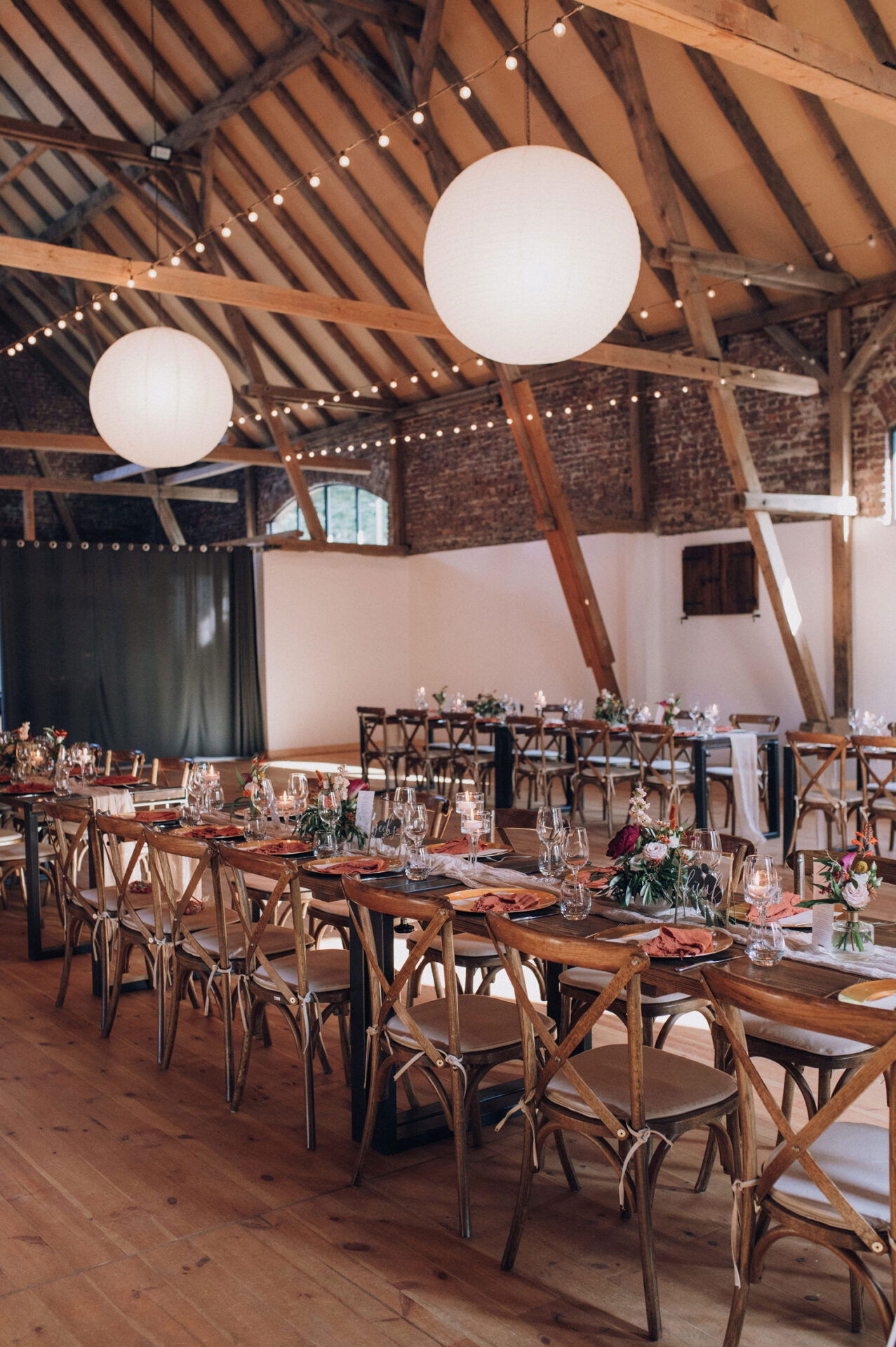 Ein rustikaler Hochzeitssaal mit Holzbalken, langen Tafeln und strahlend weißen Papierlaternen an der Decke. Die Tische sind für das Hochzeitsessen vorbereitet und mit eleganten Gläsern und Blumen geschmückt.