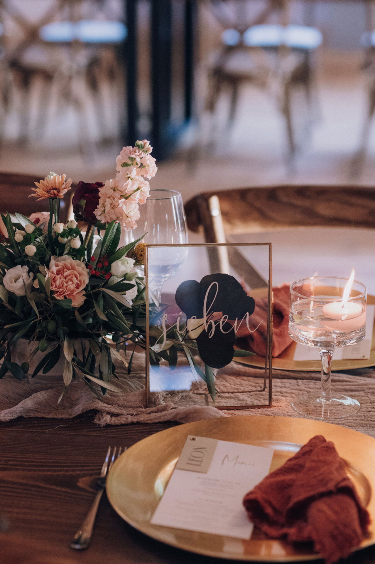 Detailaufnahme eines Tischs mit einem goldenen Teller, einer zerknitterten orangefarbenen Serviette und einem stilvollen Blumenarrangement aus rosa und roten Blüten. Eine brennende Kerze sorgt für eine warme Atmosphäre.