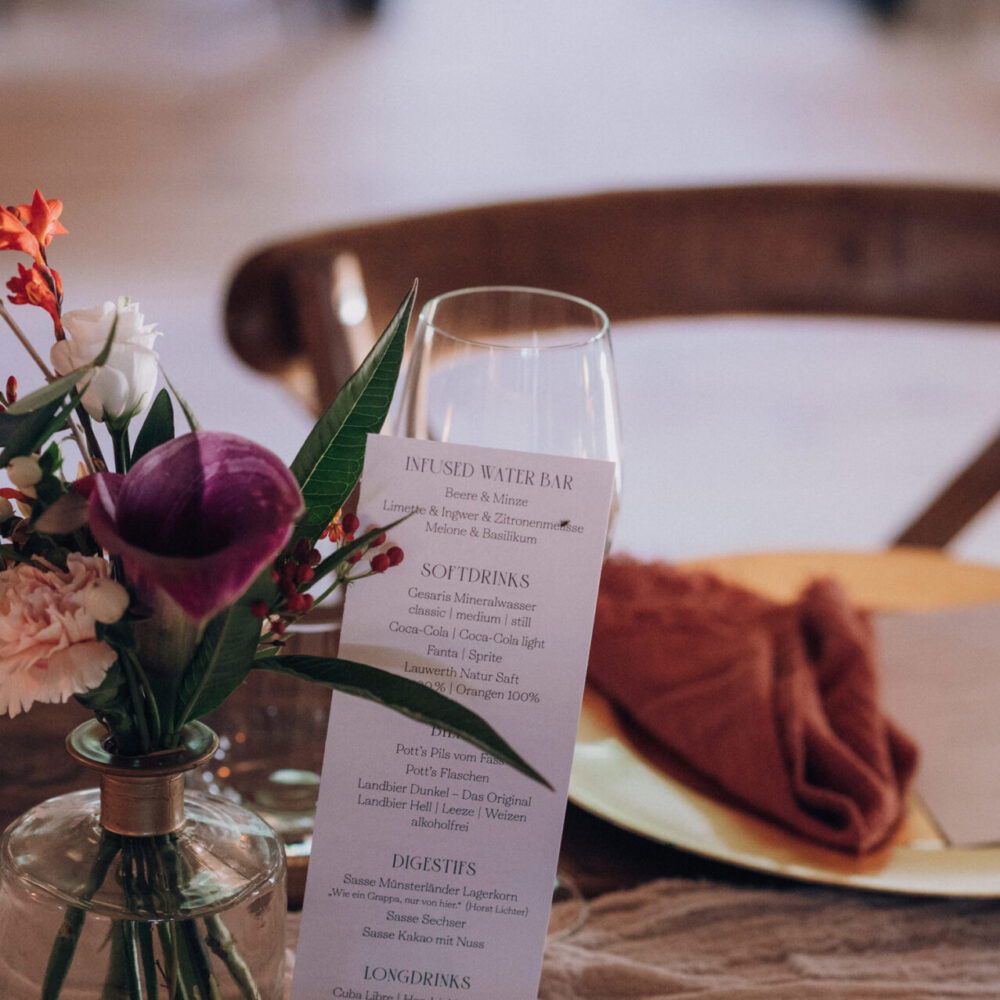 Eine stilvolle Menükarte mit verschiedenen Getränken und Speisen steht auf einem Hochzeitstisch, der durch zarte Blumendekoration und Geschirrarrangements geschmückt ist.