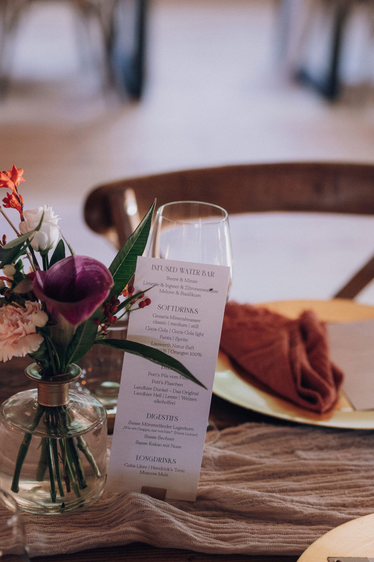 Eine stilvolle Menükarte mit verschiedenen Getränken und Speisen steht auf einem Hochzeitstisch, der durch zarte Blumendekoration und Geschirrarrangements geschmückt ist.