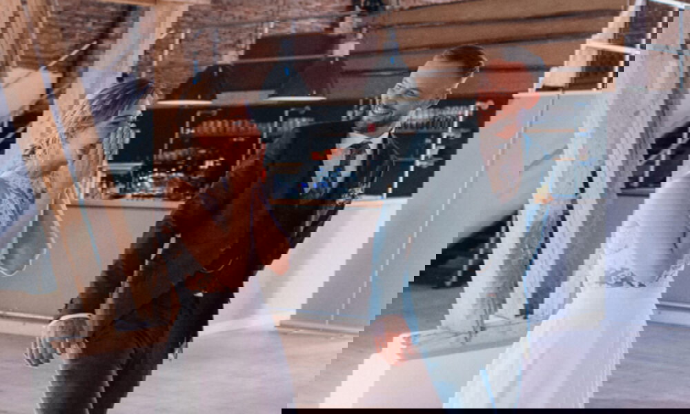 Die Braut reagiert überwältigt, als sie gemeinsam mit ihrem Bräutigam den Hochzeitsfestsaal betritt. Sie hält sich gerührt die Hände vor den Mund, während ihr Ehemann sie glücklich anlächelt. Der lichtdurchflutete Raum mit Holzelementen und einer stilvollen Bar bildet eine warme Kulisse.