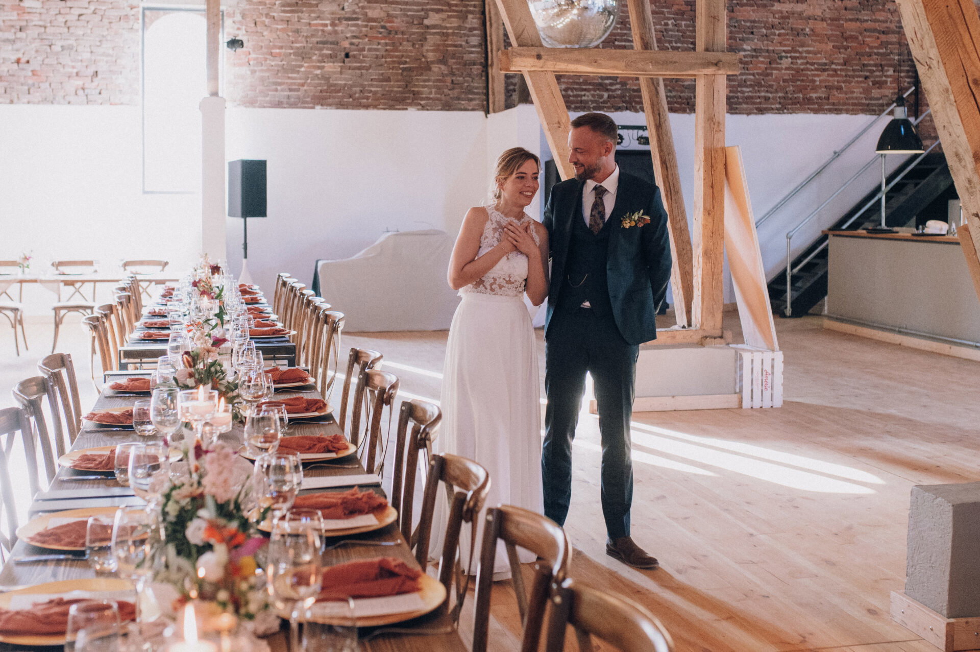 Das frisch verheiratete Paar sieht die Hochzeitslocation zum ersten Mal in voller Pracht. Die Braut wirkt überwältigt, während ihr Bräutigam sie liebevoll ansieht. Die lange, elegant gedeckte Tafel mit Blumen und Kerzen verleiht dem Raum eine feierliche Note.