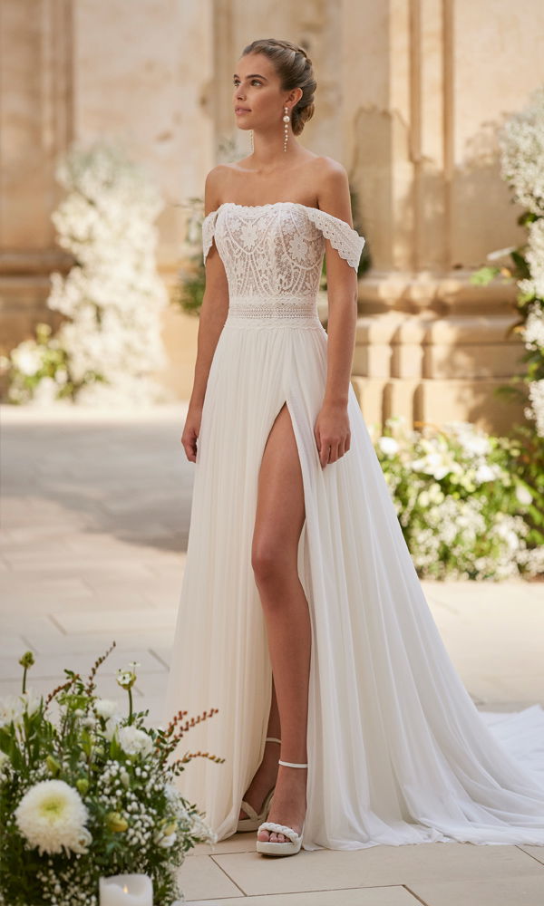 Schulterfreies Brautkleid im Boho-Stil mit verziertem Spitzenmieder und hohem Beinschlitz, umgeben von romantischem Blumenschmuck.