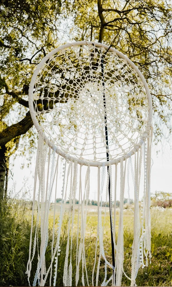 Handgefertigter Dreamcatcher aus weißer Spitze, dekorativ aufgehängt inmitten einer grünen Landschaft – ein einzigartiges Highlight für eine Outdoor-Hochzeit im Boho-Stil.