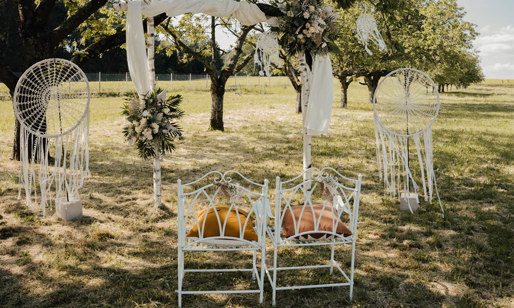 Boho-Zeremonie-Setup im Freien mit kunstvoll dekorierten weißen Stühlen, Dreamcatchern und einem mit Blumen geschmückten Altar, eingebettet in eine idyllische Baumlandschaft.