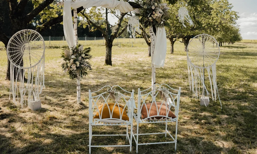 Boho-Zeremonie-Setup im Freien mit kunstvoll dekorierten weißen Stühlen, Dreamcatchern und einem mit Blumen geschmückten Altar, eingebettet in eine idyllische Baumlandschaft.