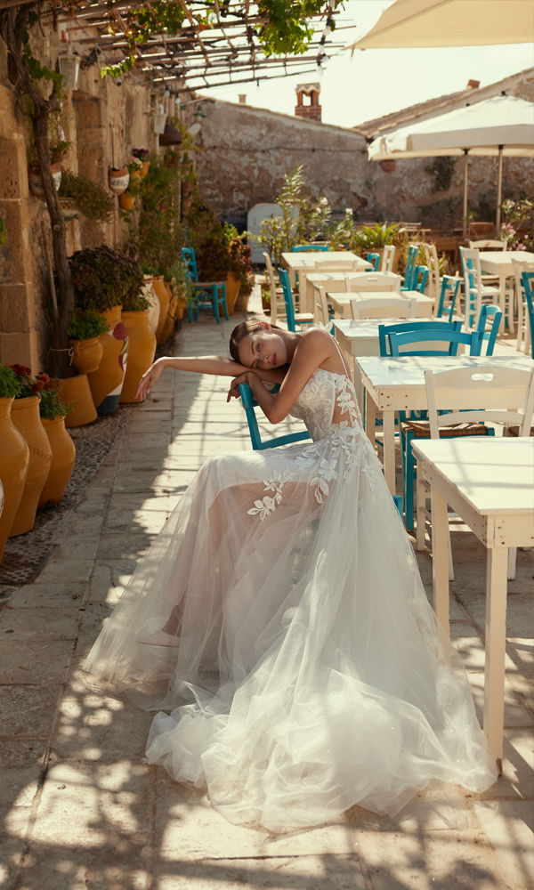 Traumhafte Braut im filigranen Tüll-Hochzeitskleid, sitzend in einem rustikalen Innenhof mit bunten Stühlen und dekorativen Vasen.