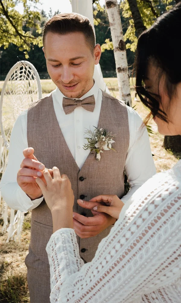 Ringtausch bei einer Boho-Hochzeit unter freiem Himmel: Der Bräutigam trägt eine Weste aus Leinen und eine Fliege, während die Braut ein Spitzenkleid trägt.