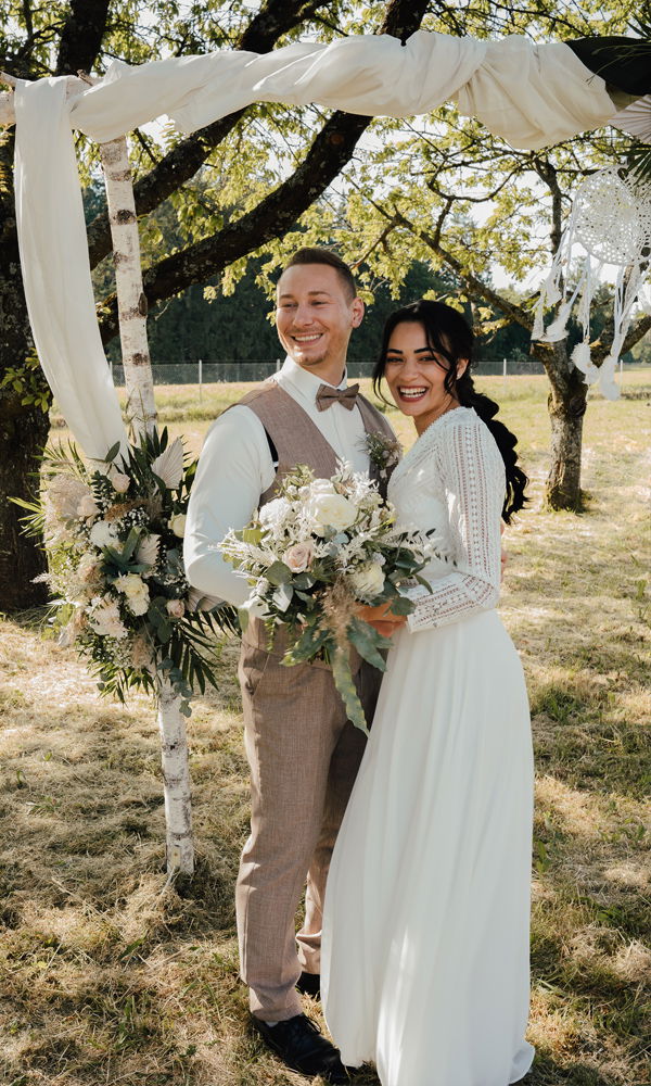 Boho-Hochzeit im Freien: Das Brautpaar lächelt unter einem mit Blumen geschmückten Bogen, umgeben von einer idyllischen Wiese und Bäumen.