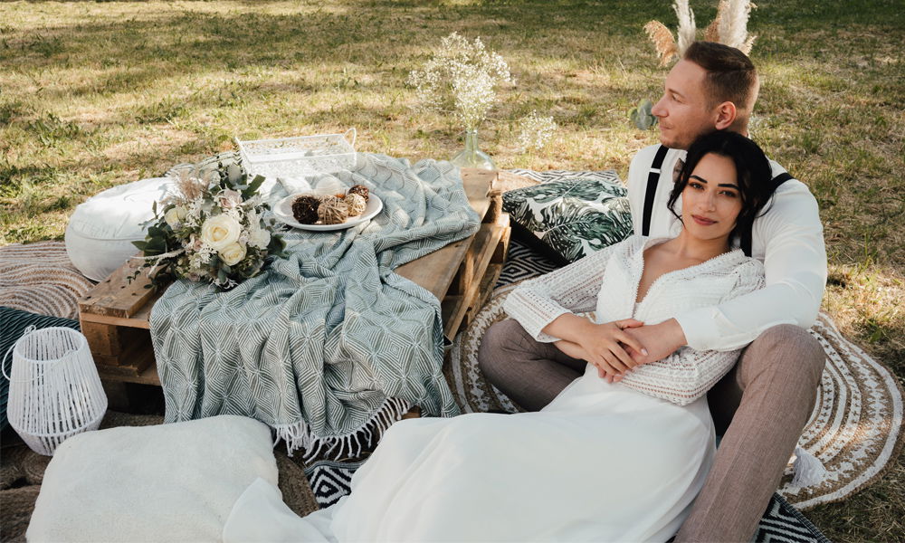 Ein verliebtes Brautpaar bei einem Boho-Picknick: Mit dekorativen Details wie Kissen, einer rustikalen Holzpalette und einem Blumenstrauß, eingebettet in die Natur.